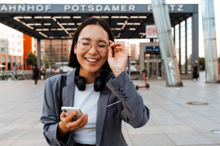 Téléchargez les photos : Jeune femme asiatique joyeuse en lunettes à l'aide d'un smartphone tout en marchant dans la rue de la ville - en image libre de droit