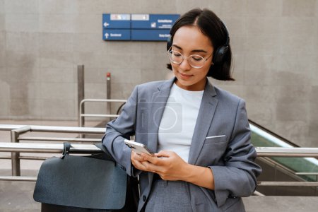 Photo for Young asian woman in headphones wearing suit using mobile phone while standing outdoors near the escalator - Royalty Free Image