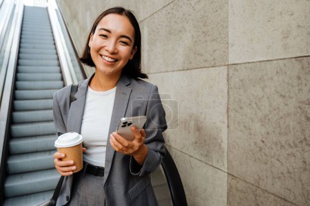 Téléchargez les photos : Young smiling asian woman holding coffee and mobile phone while standing on escalator outdoors - en image libre de droit