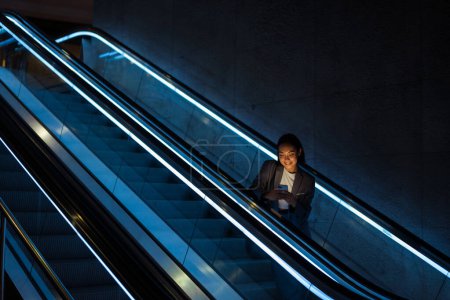 Foto de Young joyful asian woman wearing suit using mobile phone while standing on escalator indoors - Imagen libre de derechos
