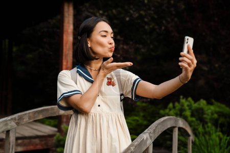 Téléchargez les photos : Jeune souriant belle asiatique fille prendre selfie sur son téléphone et souffler air baiser tout en marchant dans le parc - en image libre de droit