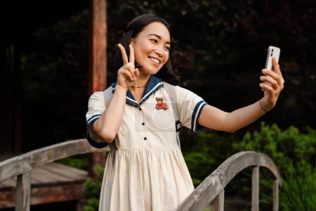 Téléchargez les photos : Young smiling beautiful asian girl taking selfie on her phone and doing peace gesture while walking in park - en image libre de droit