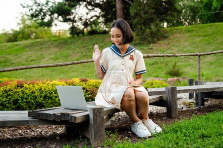Foto de Joven sonriente chica asiática usando vestido usando el ordenador portátil y saludando mientras está sentado en el banco en el parque verde - Imagen libre de derechos