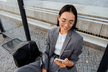 Photo for Young asian woman in glasses smiling and using mobile phone while sitting at bus station - Royalty Free Image