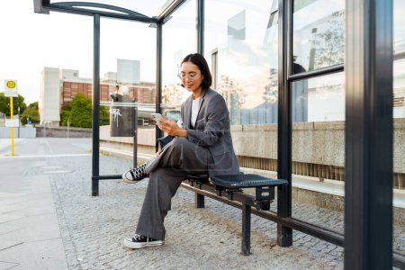 Photo for Young asian woman in glasses using mobile phone while sitting at bus station - Royalty Free Image
