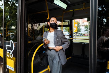 Foto de Young asian woman in medical mask with a smartphone in her hands looking aside while standing in public bus - Imagen libre de derechos