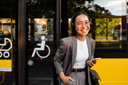 Photo for Young asian woman in headphones smiling and holding smart phone while standing near the bus - Royalty Free Image