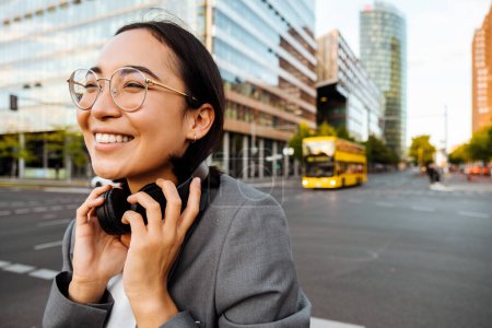 Foto de Yound asian woman in headphones smiling while standing outdoors at the city street - Imagen libre de derechos