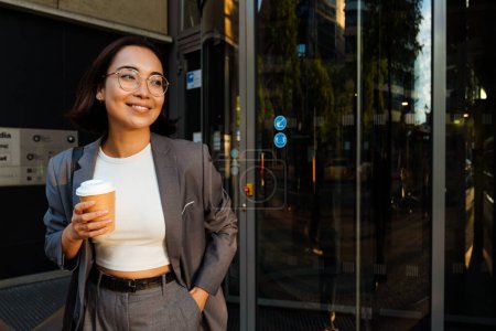 Foto de Young asian woman leaving the office building holding takeaway coffee and smiling - Imagen libre de derechos