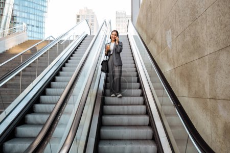Téléchargez les photos : Jeune femme asiatique parlant sur téléphone portable et tenant café à emporter tout en descendant l'escalator à l'extérieur - en image libre de droit