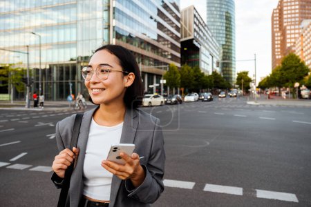 Téléchargez les photos : Jeune femme asiatique tenant téléphone portable et souriant tout en se tenant à l'extérieur à la rue de la ville - en image libre de droit