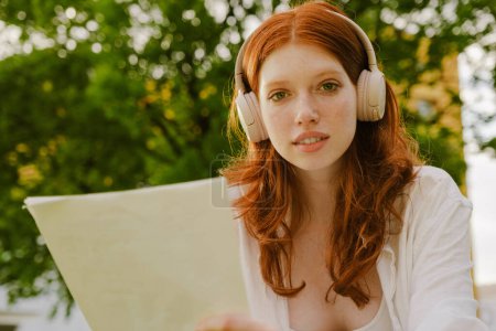 Téléchargez les photos : Portrait de jeune belle rousse en headphoenes tenant du papier et regardant un appareil photo, assis dans le parc - en image libre de droit