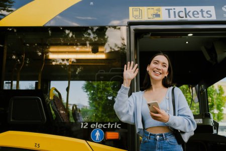 Foto de Young beautiful smiling asian girl with phone waving and looking aside, while standing near the bus outdoors - Imagen libre de derechos