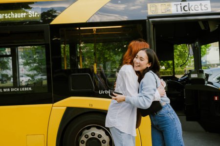 Téléchargez les photos : Jeune fille asiatique souriante mignonne étreignant sa belle amie rousse près du bus avec porte ouverte - en image libre de droit