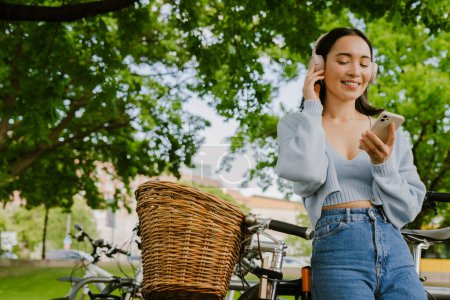 Foto de Joven hermosa sonriente chica asiática en auriculares con los ojos cerrados y el teléfono en la mano disfrutando de la música, mientras se apoya en la bicicleta de pie en el parque - Imagen libre de derechos