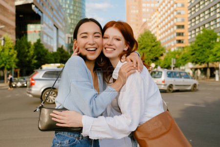 Téléchargez les photos : Two young beautiful smiling happy girls hugging each other and touching with their cheeks, looking at camera, while standing in the city on the street - en image libre de droit
