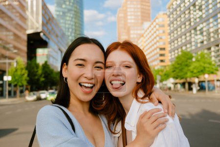 Foto de Dos jóvenes hermosas y sonrientes chicas felices abrazándose y tocando con sus mejillas una de ellas mostrando la lengua, mirando a la cámara, mientras están de pie en la ciudad en la calle - Imagen libre de derechos