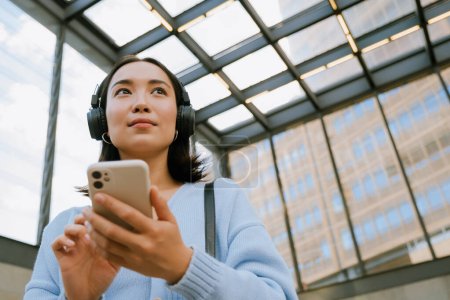 Foto de Joven mujer asiática con auriculares y el uso de teléfono móvil mientras camina en la estación de metro - Imagen libre de derechos
