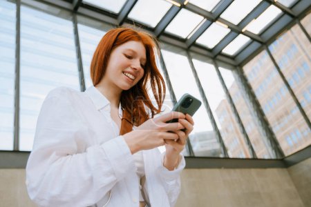 Téléchargez les photos : Jeune femme blanche souriant et utilisant un téléphone portable tout en se tenant à l'intérieur - en image libre de droit