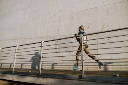 Téléchargez les photos : Asiatique mature femme en survêtement de course par mur de béton pendant l'entraînement à l'extérieur - en image libre de droit