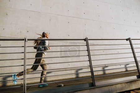 Foto de Asiática madura mujer en chándal corriendo por la pared de hormigón durante el entrenamiento al aire libre - Imagen libre de derechos