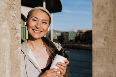 Téléchargez les photos : Asian mature woman in earphones smiling and holding water bottle during workout outdoors - en image libre de droit