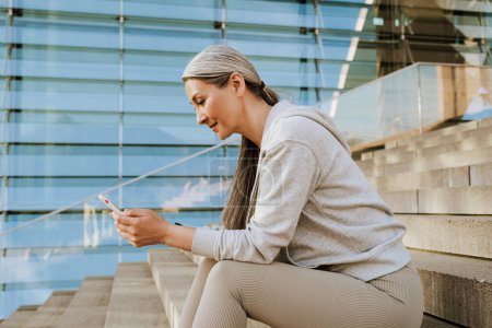 Foto de Asian mature woman in tracksuit using cellphone and sitting on stairs during workout outdoors - Imagen libre de derechos