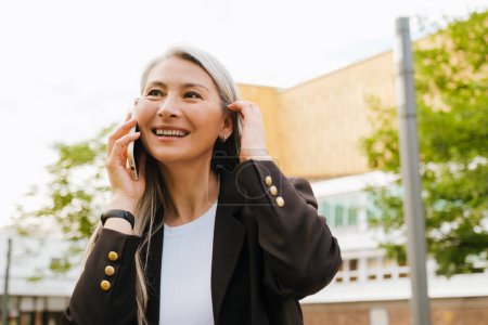 Téléchargez les photos : Grey asian woman laughing and talking on cellphone at city street - en image libre de droit