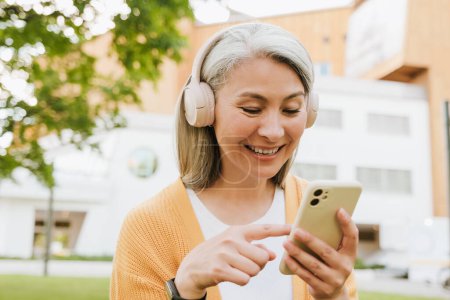Téléchargez les photos : Grey asian woman in headphones laughing and using cellphone while standing in park - en image libre de droit
