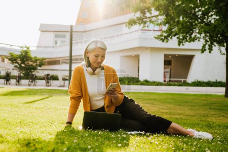 Téléchargez les photos : Grey asian woman using cellphone while working with laptop on grass at summer park - en image libre de droit