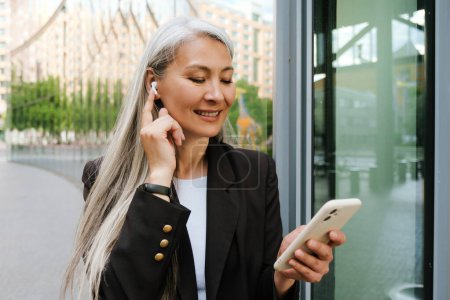 Téléchargez les photos : Grey asian woman in wireless earphones smiling and using cellphone at city street - en image libre de droit