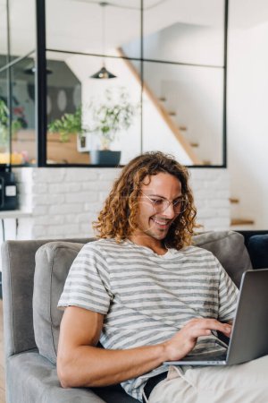 Téléchargez les photos : Jeune homme d'affaires souriant portant un t-shirt travaillant sur un ordinateur portable assis sur un canapé dans un bureau moderne - en image libre de droit