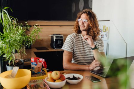 Téléchargez les photos : Young curly man sitting by table while working on laptop in office kitchen - en image libre de droit