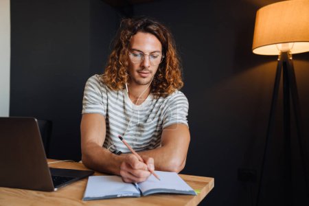 Téléchargez les photos : Young curly man using laptop and planner book while sitting by desk in office - en image libre de droit
