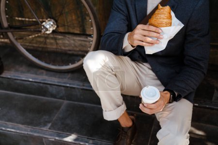 Photo for Closeup of man holding cup of coffee and croissant sitting near bicycle outdoors on city street - Royalty Free Image
