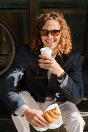 Photo for Ginger man in sunglasses holding croissant and cup of coffee while sitting outdoors on city street - Royalty Free Image