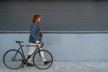 Photo for Ginger european man walking with bicycle near grey wall outdoors on city street - Royalty Free Image