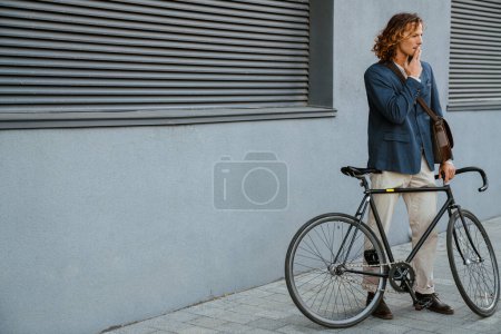 Photo for Young ginger long-haired man smoking cigarette while standing with bicycle near grey wall outdoors - Royalty Free Image