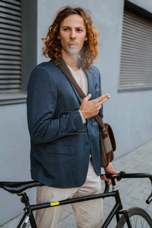 Photo for Young ginger long-haired man smoking cigarette while standing with bicycle near grey wall outdoors - Royalty Free Image
