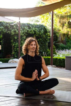 Foto de Joven hombre rizado sonriendo mientras está sentado en el suelo de madera en el estudio de yoga al aire libre - Imagen libre de derechos