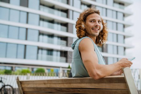 Photo for Young handsome smiling sporty long-haired man in headphones with phone looking aside, while sitting on the bench outdoors - Royalty Free Image