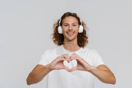 Photo for Ginger european man in headphones smiling and doing heart-shape gesture isolated over white background - Royalty Free Image