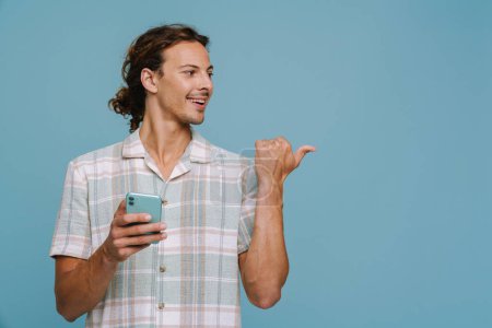 Foto de Hombre europeo jengibre señalando con el dedo a un lado y utilizando el teléfono celular aislado sobre fondo azul - Imagen libre de derechos