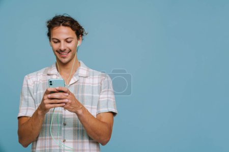 Photo for Ginger european man in earphones smiling and using cellphone isolated over blue background - Royalty Free Image