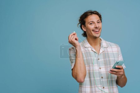 Photo for Ginger european man in earphones smiling and using cellphone isolated over blue background - Royalty Free Image