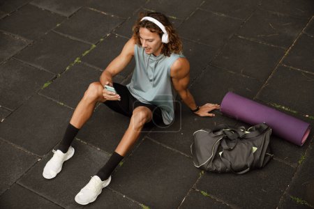Téléchargez les photos : Jeune homme athlétique aux cheveux longs dans des écouteurs avec téléphone assis sur le sol avec tapis et sac sur le terrain de sport à l'extérieur - en image libre de droit