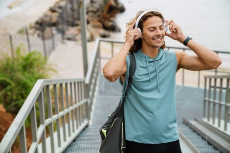 Photo for Portrait of young long-haired athletic smiling man in headphones with closed eyes enjoying musi? , while walking up the stairs outdoors - Royalty Free Image