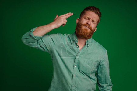 Photo for Adult handsome stylish bearded serious man in green shirt doing suicide gesture with closed eyes , while standing over isolated green background - Royalty Free Image