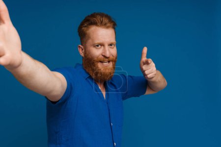 Foto de Selfie of adult handsome redhead bearded smiling man doing gun gesture with fingers and looking at camera, while standing over isolated blue background - Imagen libre de derechos