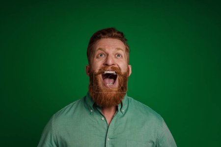Téléchargez les photos : Portrait of adult handsome stylish bearded shouting man in green shirt looking upwards , while standing over isolated green background - en image libre de droit